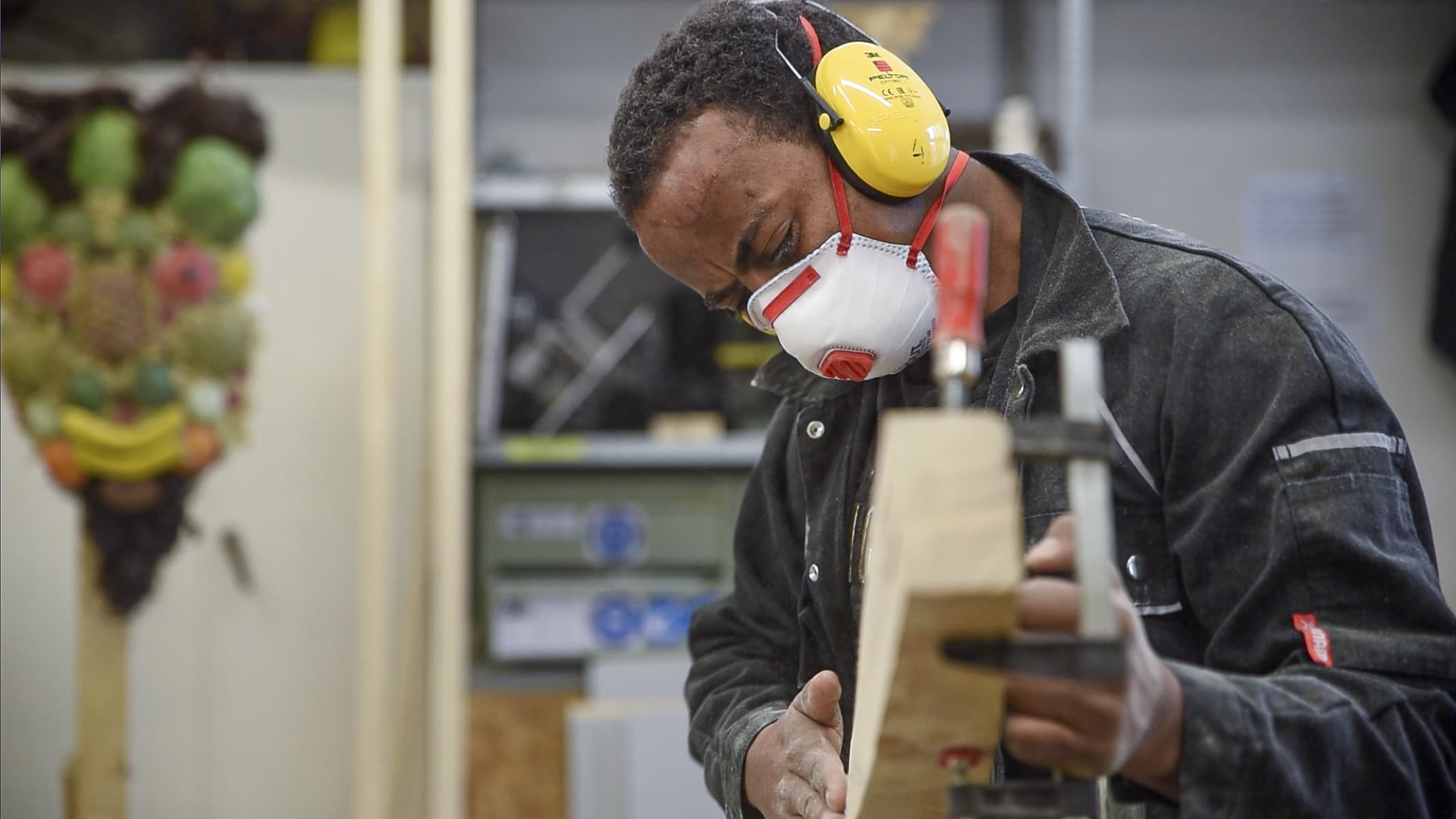 Eine Person trägt Mund- und Hörschutz. Sie hält ein mittelgroßes Stück Holz in der einen Hand und fühlt mit der anderen Hand, ob die Kanten gerade geschnitten sind.