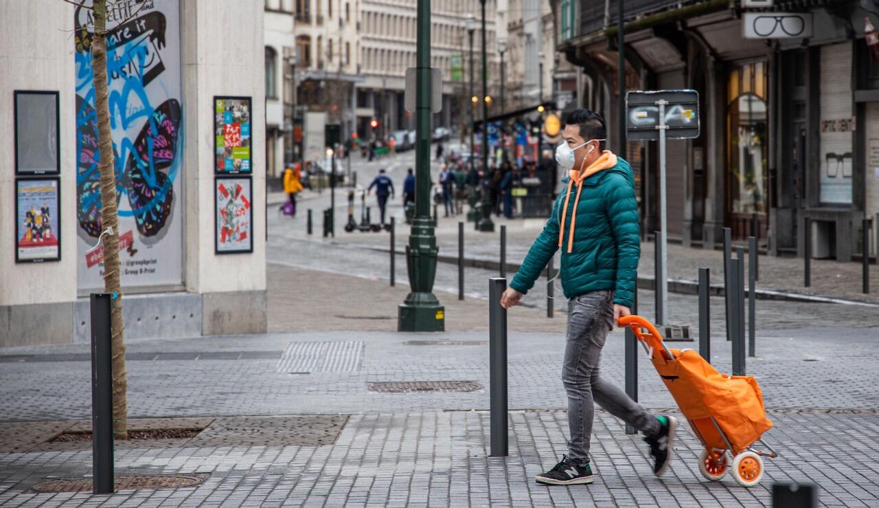 Das Bild zeigt einen Mann auf einer Straße in einer großen Stadt. Er zieht hinter sich einen orangen Einkaufstrolley und trägt eine FFP-Schutzmaske.