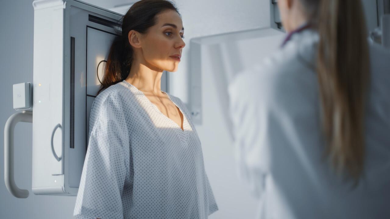 Das Bild zeigt eine Frau in einem Arztkittel. Sie steht vor einem Röntgenaufnahmegerät. Rechts im Bild ist eine Arzthelferin zu sehen.