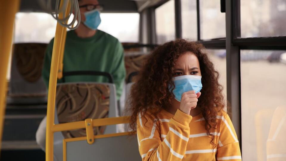 Das Bild zeigt eine Frau im Bus. Sie trägt eine medizinische Mundschutzmaske und hält sich eine Hand vor den Mund.
