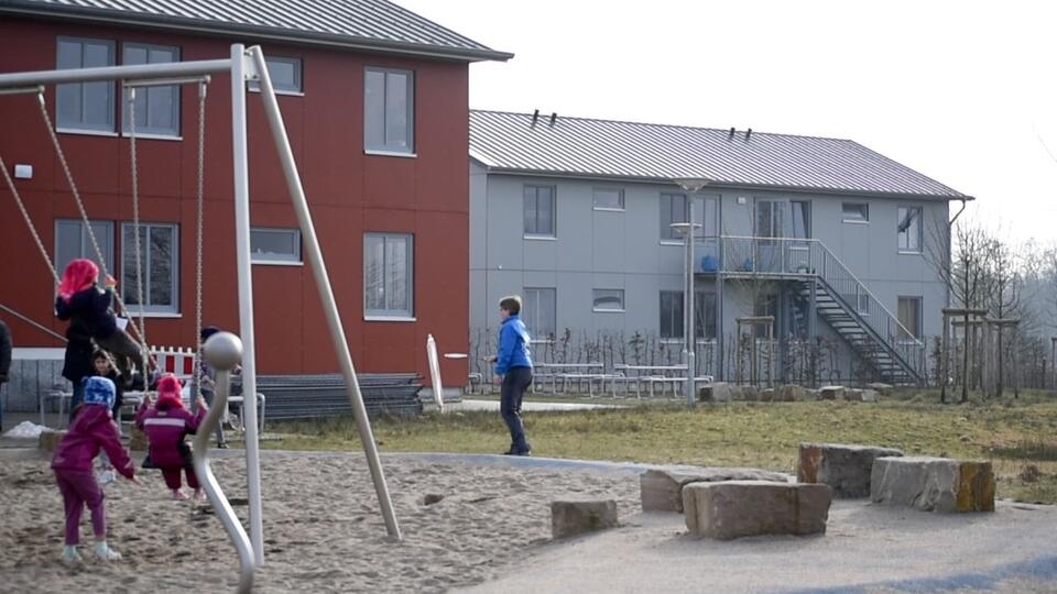 Das Bild zeigt einen Spielplatz vor zwei Häusern. Auf dem Spielplatz schaukeln Kinder. 