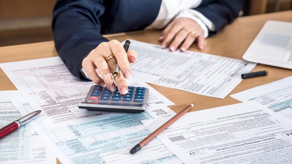 Das Bild zeigt einen Schreibtisch, auf dem einzelne Seiten einer Steuererklärung liegen. Am Schreibtisch sitzt ein Mann, der die Steuererklärun mit Hilfe eines Taschenrechners und Stift ausfüllt.