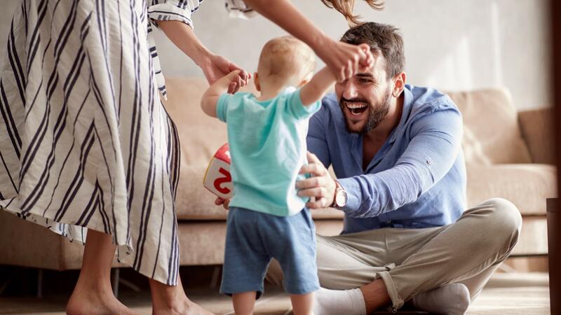 Das Bild zeigt eine Familie im Wohnzimmer. Der Vater öffnet seine Hände, um sein Kleinkind in die Arme zu nehmen. 