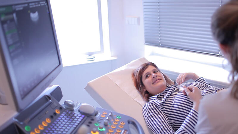 Das Bild zeigt eine schwangere Frau in einer Arztpraxis. Sie liegt auf einer Untersuchungsliege. Eine Ärztin untersucht ihren Bauch mit einem Ultraschallgerät. Links im Bild sieht man den Bildschirm des Ultraschallgeräts. 