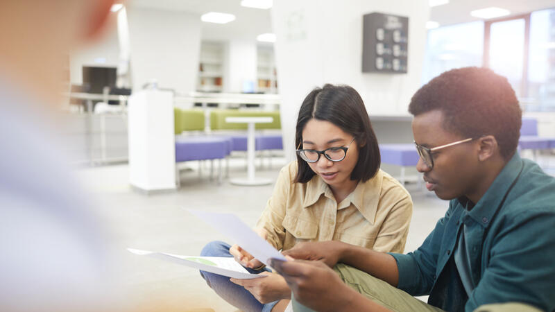 Das Bild zeigt eine Studentin und einen Studenten. Sie sitzen zusammen und schauen sich Formulare an. Im hintergrund sieht man eine Sitzugruppe mit Tischen und Bänken.